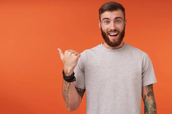 Happy pretty bearded tattooed man with trendy haircut looking at camera with wide smile and rounding his blue eyes, pointing aside with raised thumb while posing over orange background — Stock Photo, Image