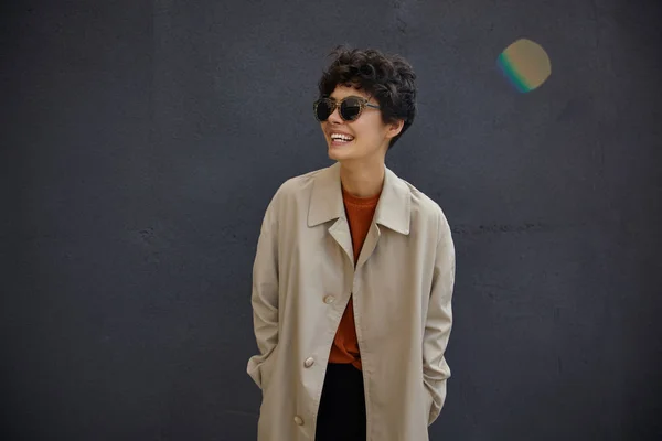 Outdoor shot of young pretty woman with short curly hair standing over black urban wall while waiting for her friends, looking aside joyfully with wide happy smile, wearing trendy outfit