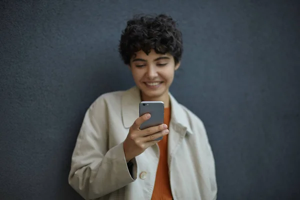 Outdoor shot van jonge donkerharige krullende vrouw met kort kapsel poseren tegen zwarte stedelijke muur, houden mobiele telefoon in opgeheven hand en kijken positief op het scherm — Stockfoto