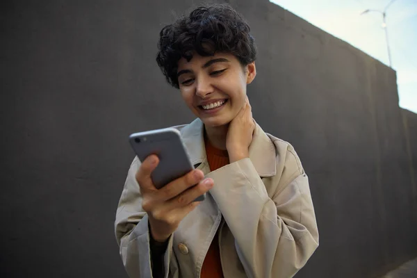 Close-up van vrolijke aantrekkelijke jonge brunette vrouw met kort krullend haar houden mobiele telefoon in de hand en houd haar nek, kijken naar het scherm gelukkig met brede glimlach — Stockfoto