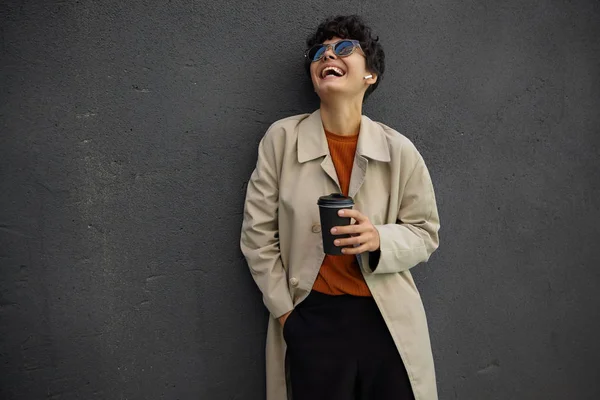 Vrolijke aantrekkelijke jonge krullende vrouw met donker kort haar vrolijk lachen en gooien haar hoofd terug, staande over stedelijke omgeving tijdens het drinken van koffie voor werkdag — Stockfoto