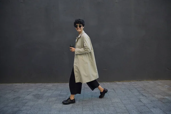 Positieve krullende donkerharige zakenvrouw met kort kapsel wandelen over stedelijke omgeving met zwarte papieren beker, gaan lunchen buiten kantoor, het dragen van trendy kleding en stijlvolle zonnebril — Stockfoto
