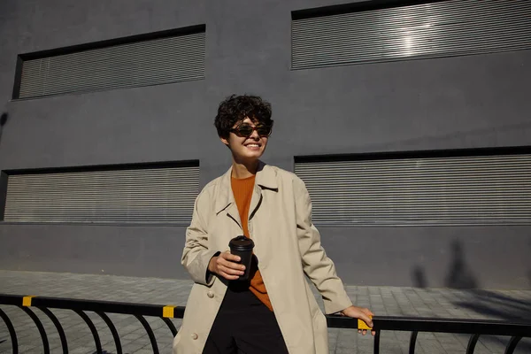 Lifestyle fashion foto van jonge donkerharige stijlvolle hipster vrouw met kort kapsel wandelen over stedelijke omgeving in beige trench jas en foxy trui, op zoek opzij met oprechte glimlach — Stockfoto