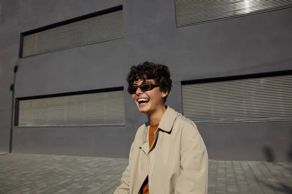 Outdoor shot van vrij gelukkig krullend brunette vrouw in vintage zonnebril en beige trench jas poseren over de stad milieu tijdens het weekend, vrolijk lachen en tonen haar perfecte witte tanden — Stockfoto
