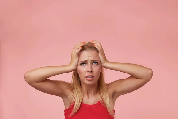 Retrato interior de la joven rubia estresada en camisa roja, sosteniendo las sienes con las manos, mirando hacia arriba y frunciendo el ceño, teniendo mal día, aislada sobre fondo rosa — Foto de Stock