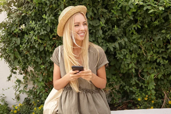 Retrato al aire libre de una linda joven rubia con un vestido de lino informal sobre un jardín verde, manteniendo el teléfono inteligente en las manos y mirando a un lado con una amplia sonrisa — Foto de Stock