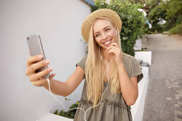 Joven mujer rubia feliz escuchando música mientras hace selfie con su teléfono móvil, vistiendo vestido de lino casual y sombrero de paja, luciendo alegre y complacido — Foto de Stock