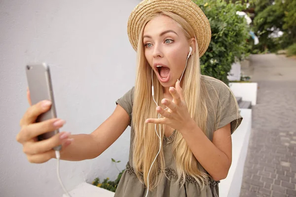 Sorprendido atractiva dama rubia en sombrero de paja y vestido romántico hablando con alguien por chat de vídeo, caminando por la calle verde en buen día brillante — Foto de Stock