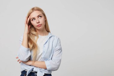 Beyaz arka plan üzerinde poz veren, uzun saçlarını elleriyle düzleştiren ve hayret verici bir şekilde bir kenara bakan kafası karışmış genç sarışın bir kadının stüdyo portresi.