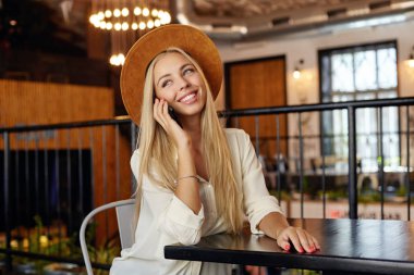 Güzel, uzun saçlı, beyaz tişörtlü, kahverengi şapkalı bir kadın yemek molasında restoranda otururken telefonda hoş sohbetler ediyor.
