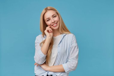 Mavi tişörtlü, mavi tişörtlü, gri tişörtlü, mavi arka planda poz veren çekici bir şekilde kameraya bakan ve elini kaldırmış dface 'e dokunan sarışın kadın.