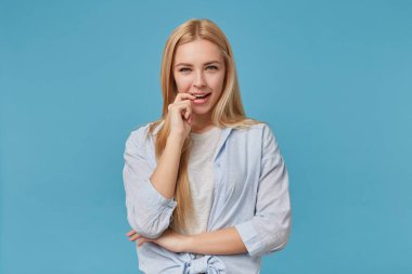 Flört eden, uzun saçlı mavi arka planda dikilen mavi gömlekli, gri tişörtlü, şaşı ve ısıran işaret parmağıyla kameraya bakan genç bayanın portresi.