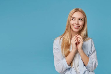 Sıradan saç stili olan genç sarışın bir kadının stüdyo fotoğrafı katlanmış elleri kaldırıyor, mavi arka planda poz veriyor, neşeli bir gülümsemeyle yan yana bakıyor.