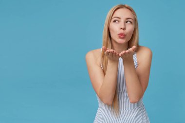 Sarışın, uzun saçlı, mavi arka planda poz veren, romantik bir yaz elbisesi giyen, güzel bakışlı ve kameraya hava öpücüğü veren bir kadının yatay görüntüsü.