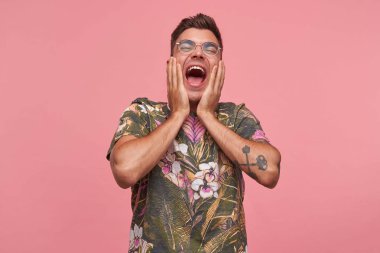 Yakışıklı, kısa saçlı, çiçekli tişörtlü, pembe arka planda duran, avuçlarını yüzüne doğru kaldırmış, kafasının arkasına doğru fırlatılan bir erkek.