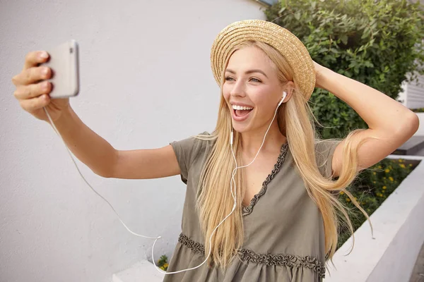 Mooie jonge vrouw met lang blond haar met haar strohoed en het maken van zelfportret op mobiele camera, brede glimlach en op zoek gelukkig — Stockfoto