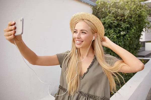 Retrato al aire libre de una joven rubia feliz haciendo selfie con su teléfono inteligente, usando un vestido de lino casual y un sombrero de paja, luciendo feliz y alegre — Foto de Stock