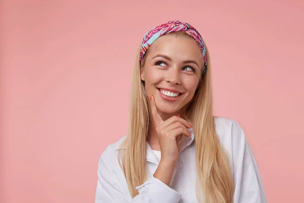 Estúdio tiro de encantadora mulher bonita, vestindo headband e camisa branca, tocando seu queixo e olhando para longe com amplo sorriso, posando sobre o fundo rosa — Fotografia de Stock