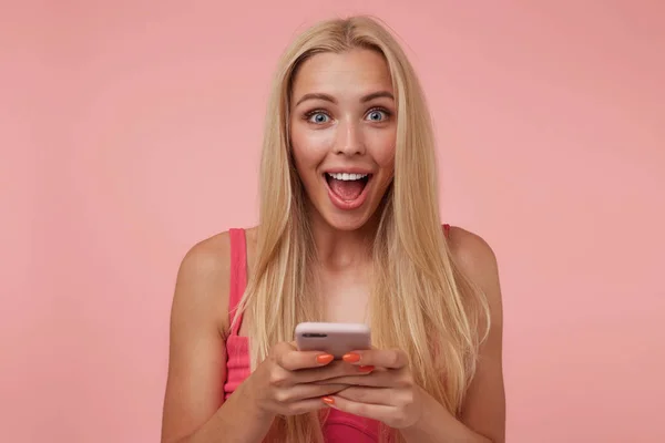 Retrato de mujer joven y bonita sorprendida con el pelo largo y rubio de pie aislado sobre el fondo rosa, sosteniendo el teléfono móvil y mirando a la cámara — Foto de Stock