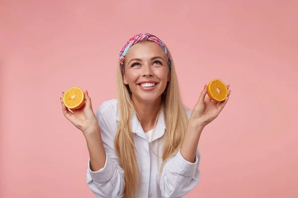 Hoş görünümlü sarışın bir kadının mutlu bir şekilde yukarı baktığı, parlak geniş bir gülümsemesi olan, pembe arka planda duran, ellerinde portakal parçalarıyla çekilmiş bir fotoğraf. — Stok fotoğraf