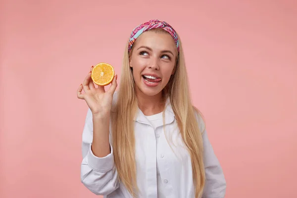 Linda jovem loira posando sobre fundo rosa com laranja na mão, puxando a língua e olhando para o lado, vestindo roupas casuais — Fotografia de Stock
