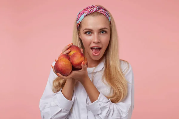 Mulher jovem atraente excitado com pêssegos em suas mãos olhando para a câmera com a boca aberta larga, vestindo headband colorido e camisa branca, isolado sobre fundo rosa — Fotografia de Stock