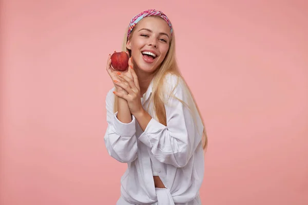 Jovem linda mulher loira com manicure laranja em camisa branca olhando para câmera e segurando pêssego perto da bochecha, sendo positivo e feliz, isolado sobre fundo rosa — Fotografia de Stock