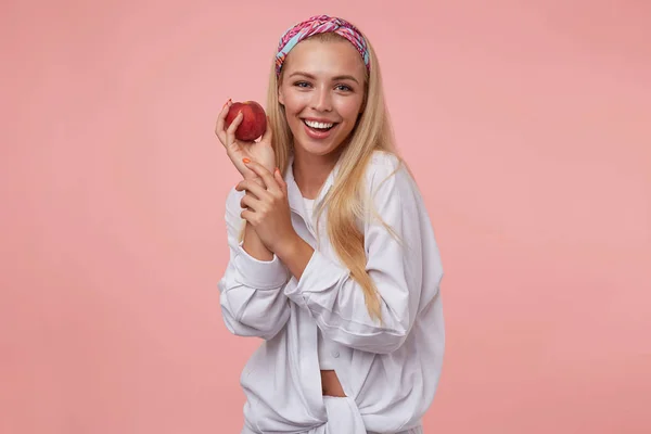 Foto interior da mulher bonita alegre sorrindo amplamente e posando com pêssego na mão sobre o fundo rosa, vestindo roupas casuais — Fotografia de Stock