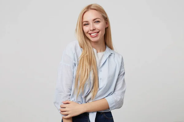 Retrato interior de jovem loira atraente mulher de cabelos longos sorrindo para câmera sinceramente, posando sobre fundo branco, vestindo jeans e camisa azul — Fotografia de Stock