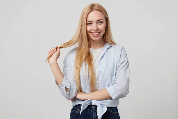 Retrato interno de jovem loira de aparência agradável sorrindo para a câmera sobre fundo branco, segurando um fio de cabelo, vestindo roupas casuais — Fotografia de Stock