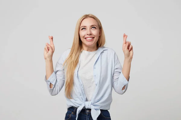 Indoor shot van een lachend jong meisje met lang blond haar in blauw shirt en jeans, staande over witte achtergrond, vingers gekruist voor geluk en naar boven kijkend — Stockfoto