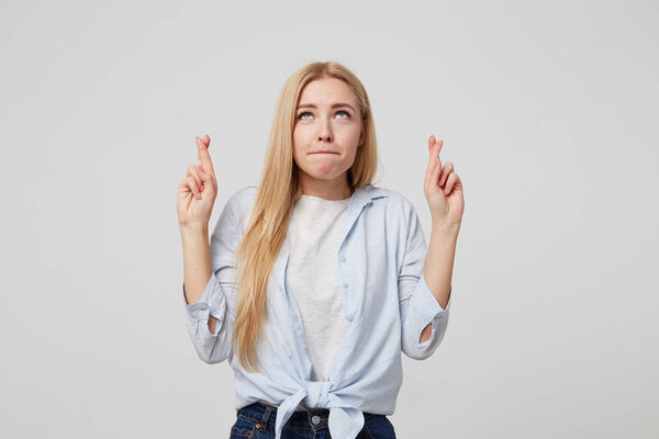 Portrait of attractive young woman in blue shirt and jeans crossing fingers with great hope for better, wishing for good luck, posing over white background