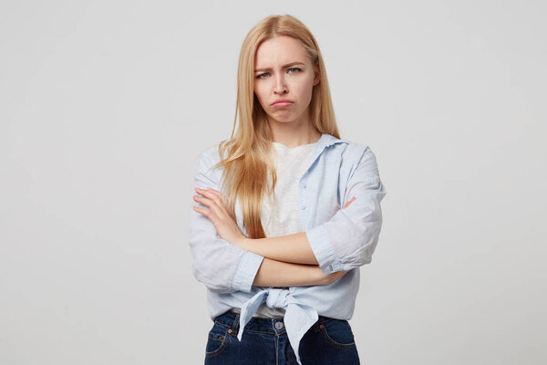 Indoor portrait of charming young blonde female standing over white background with crossed arms, looking offended and upset, frowning and pouting her lips