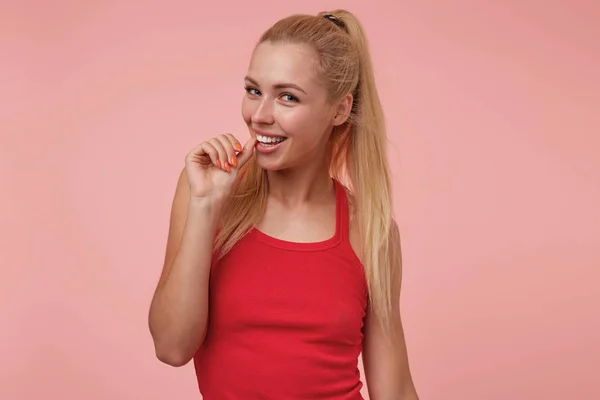 Estúdio tiro de flerte jovem bonita senhora com longos cabelos loiros em camisa vermelha, sorrindo brincando e mantendo a mão perto da boca, isolado sobre fundo rosa — Fotografia de Stock