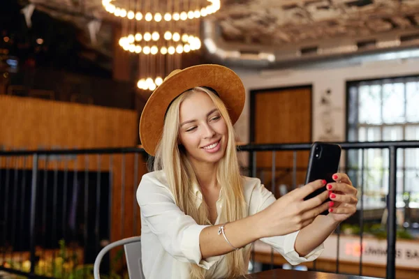 Hermosa joven rubia de pelo largo con sombrero y camisa blanca, posando sobre el interior de la cafetería, sonriendo ampliamente a la cámara mientras hace selfie con su teléfono inteligente — Foto de Stock
