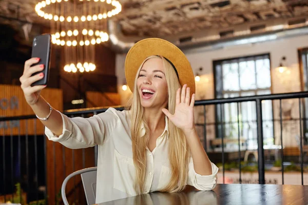 Atractiva joven rubia de pelo largo sentada en la mesa de la cafetería mientras almorza, haciendo una foto de sí misma con su teléfono inteligente, levantando la palma en gesto de saludo y sonriendo ampliamente — Foto de Stock