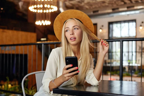 Beautiful blonde pretty woman in hat posing over restaurant during lunch break with mobile phone in hand, looking aside thoughtfully and touching her hair