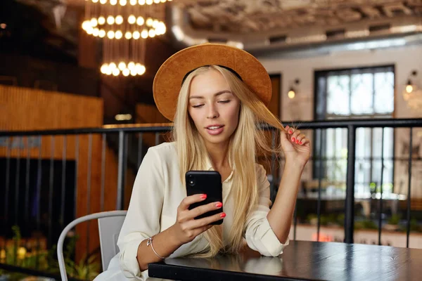 Foto interior de una guapa joven rubia de pelo largo sentada en la mesa sobre el interior de la cafetería, manteniendo el teléfono inteligente en la mano y mirando la pantalla con una sonrisa suave — Foto de Stock