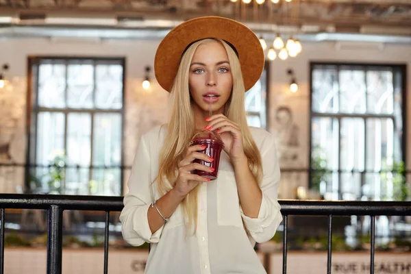 Indoor shot de hermosa mujer rubia de pelo largo con ojos azules posando sobre el interior del restaurante, bebiendo limonada con paja, usando sombrero y camisa — Foto de Stock