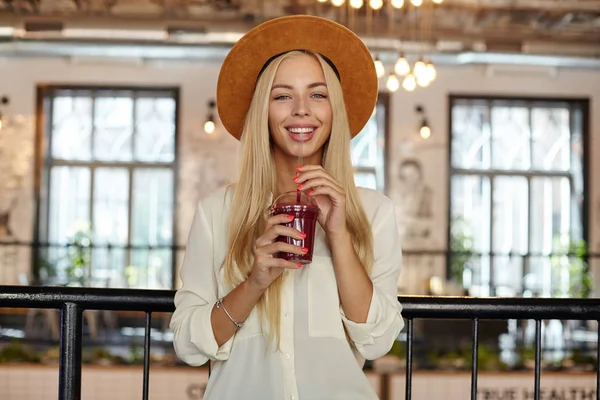 Alegre hermosa joven hembra con el pelo largo y rubio mirando a la cámara felizmente mientras posa sobre el interior de la cafetería, sosteniendo la taza de limonada en las manos — Foto de Stock