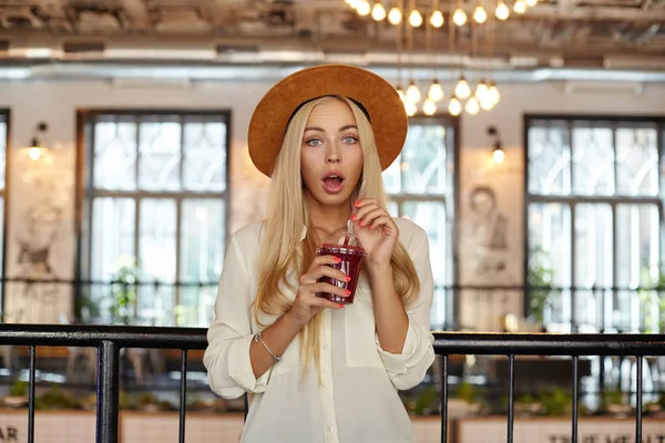 Mujer rubia joven sorprendida en sombrero ancho posando sobre el interior de la cafetería, mirando a la cámara con la boca abierta y sosteniendo la taza de limonada en sus manos — Foto de Stock