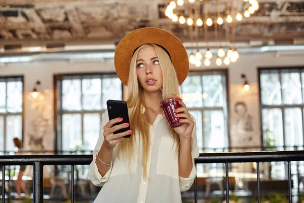 Atractiva joven rubia bebiendo batido con paja mientras espera su pedido en la cafetería, sosteniendo el teléfono inteligente en la mano y mirando a un lado con interés — Foto de Stock