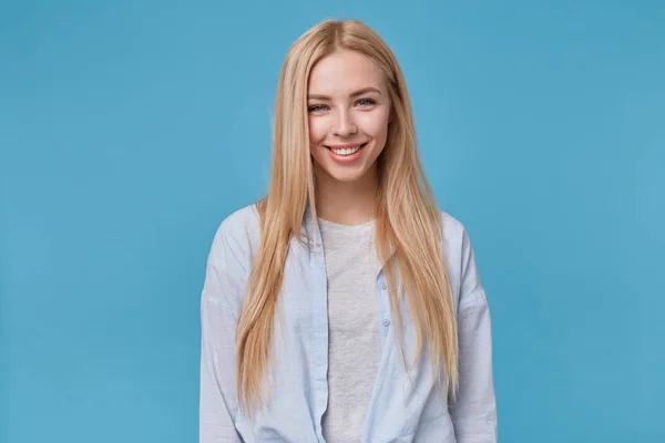 Retrato de jovem mulher loira com penteado casual posando sobre fundo azul, vestindo camisa azul e camiseta cinza, mostrando emoções positivas após receber elogio agradável — Fotografia de Stock