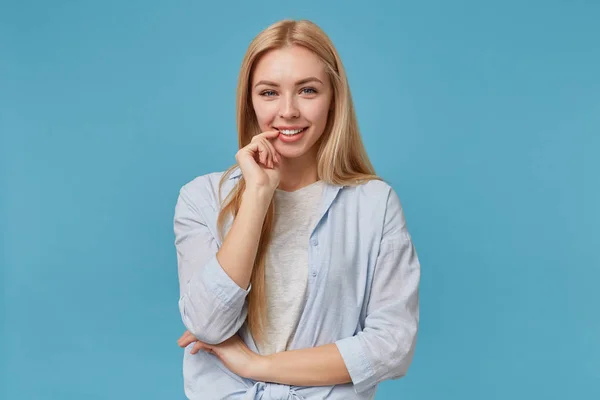 Sorrindo positivo fêmea loira com aparência atraente e cabelos longos posando sobre fundo azul, segurando o dedo indicador na parte inferior do lábio e olhando para a câmera alegremente, vestindo roupas casuais — Fotografia de Stock