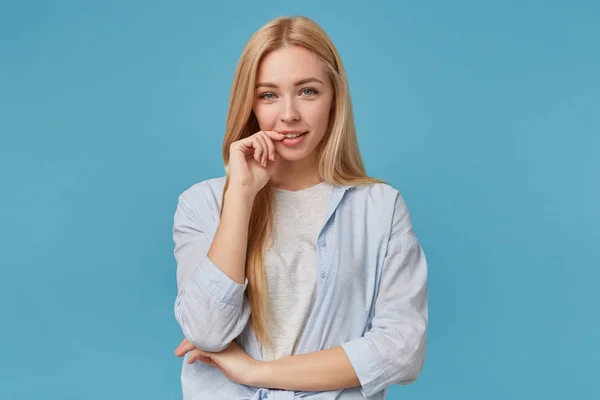 Foto horizontal de mulher loira de cabelos longos positiva em roupas casuais posando sobre fundo azul, mantendo o dedo indicador na parte inferior do lábio e sorrindo para a câmera sinceramente — Fotografia de Stock