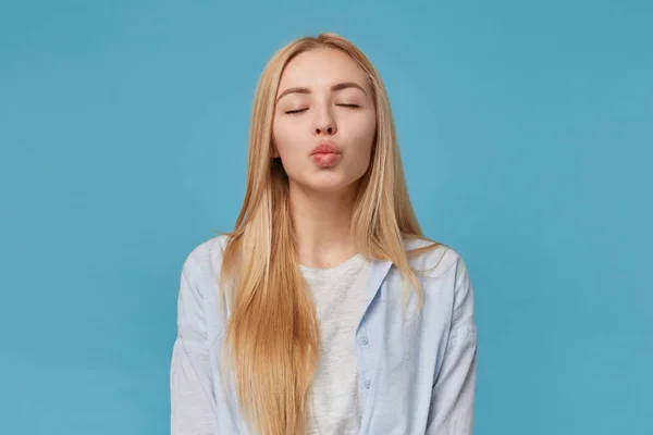 Estúdio foto de jovem linda fêmea com cabelo longo loiro vestindo roupas casuais, posando sobre fundo azul com olhos fechados e lábios dobráveis no beijo ar — Fotografia de Stock
