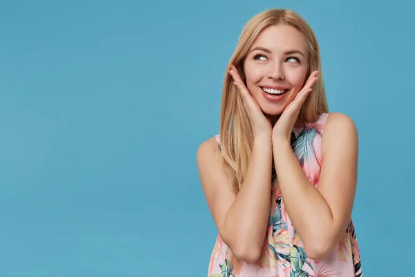 Studio colpo di bella giovane donna bionda con i capelli lunghi indossa abito fiorito, guardando sognante verso l'alto e sorridente felicemente, isolato su sfondo blu con palme sollevate — Foto Stock