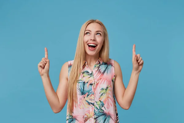 Indoor shot van positieve blonde vrouw met lang haar naar boven wijzend met wijsvingers, staande over blauwe achtergrond in bloeiende jurk, vrolijk glimlachend en haar witte perfecte tanden demonstrerend — Stockfoto