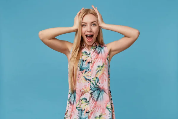 Retrato de mulher loira jovem espantada com penteado casual posando sobre fundo azul, mantendo as palmas na cabeça e olhando alegremente para a câmera com a boca larga aberta — Fotografia de Stock