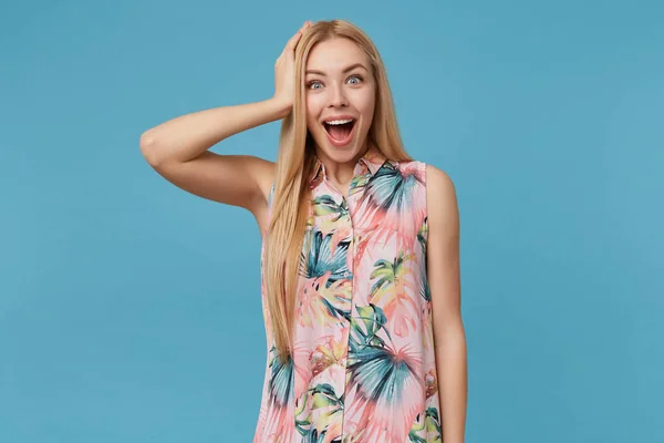 Surpreendida mulher loira bonita com cabelos longos posando sobre fundo azul em vestido romântico com estampa floral, levantando a mão feliz para a cabeça enquanto sorri amplamente para a câmera com olhos abertos — Fotografia de Stock